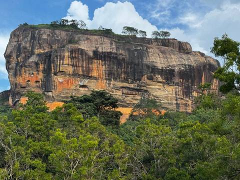       Massive Sigiriya rock fortress rises above dense jungle under a partly cloudy sky.
  