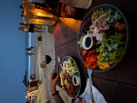       Dinner plates of colorful salads and wine on a beachside table at dusk with swimmers on the sand.
  