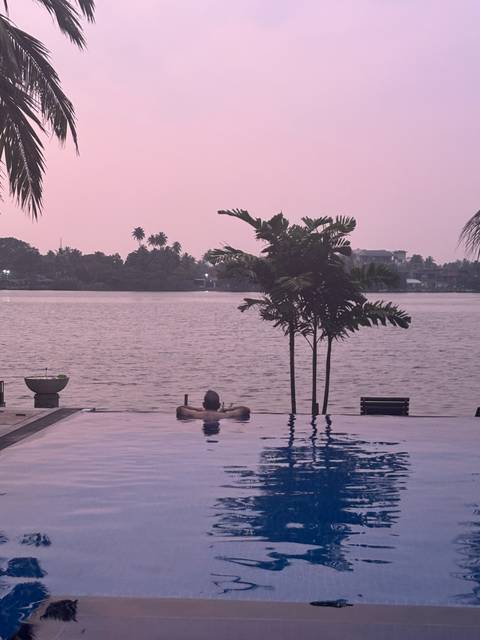       Person relaxing in an infinity pool facing a calm lagoon at pastel pink sunset with palms silhouetted
  