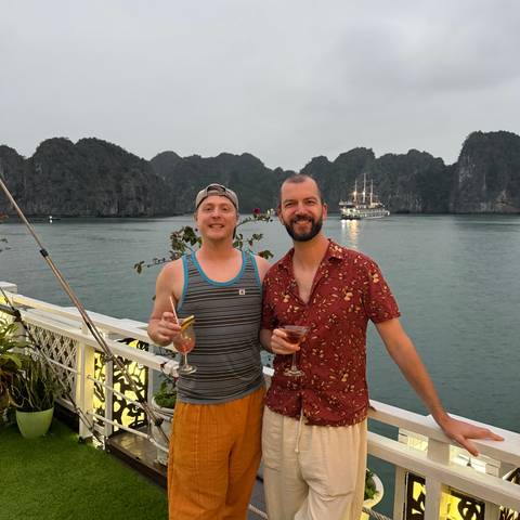       Two friends toast cocktails on a deck with limestone karst islands and a cruise ship in the background.
  