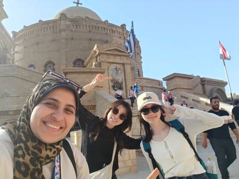       Smiling visitors in front of an ornate stone church and waving national flags in Cairo.
  