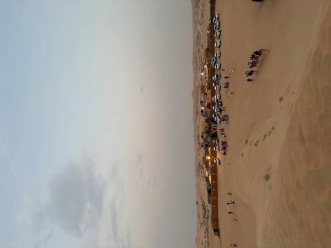      Wide desert scene of evening camp, 4x4s and camel caravan under fading light.
  