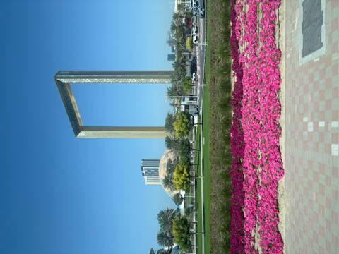       Towering golden Dubai Frame rising above manicured park with bright pink flowers.
  