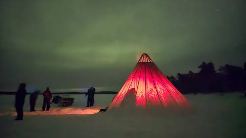       Red canvas tepee glowing from within on snowy ground as silhouetted people observe the night sky.
  