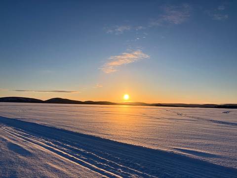       Golden sun setting over a vast frozen lake with distant low hills and snowmobile tracks.
  