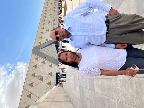       Mature couple posing outside the modern Grand Egyptian Museum with geometric facade under blue sky.
  
