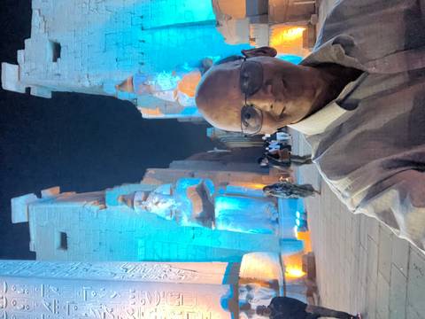       Selfie of a man at night with colossal pharaoh statues of Luxor Temple illuminated in blue lights behind.
  