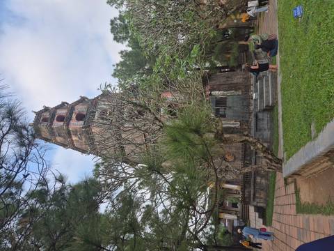       Tall ancient Thien Mu pagoda in Hue framed by pine trees with two visitors seated nearby.
  
