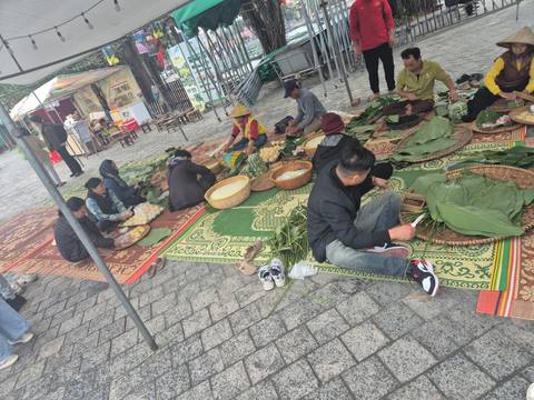       Local people sitting on mats preparing traditional food parcels with banana leaves in an outdoor market setting.
  