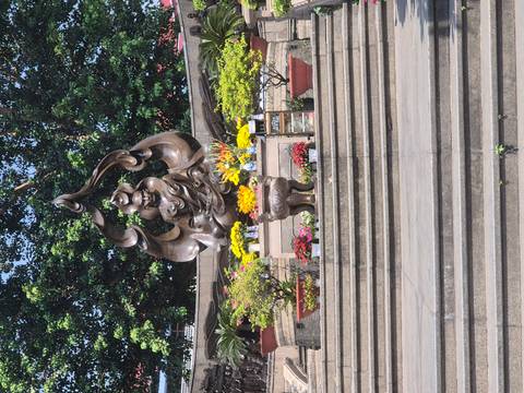      Outdoor bronze statue surrounded by colourful flower offerings and greenery on temple steps.
  