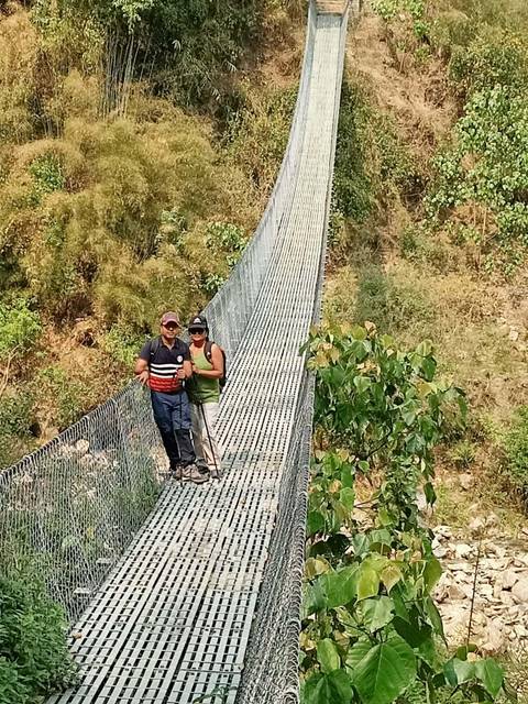       Two trekkers stand on a long metal suspension bridge spanning a lush gorge in rural Nepal.
  