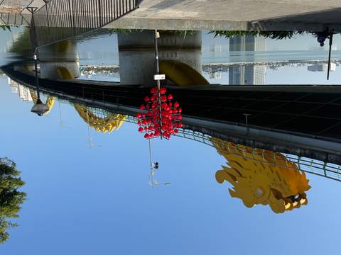       Golden dragon-shaped bridge arching over a calm river in bright morning light with a heart-shaped lantern sculpture in front.
  