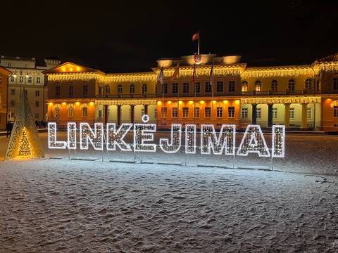       Illuminated "LINKĖJIMAI" light sign shines on a snowy square against a backdrop of a lit classical building.
  