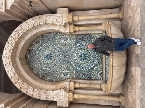       Traveler poses at ornate zellige mosaic fountain within Hassan II Mosque complex.
  