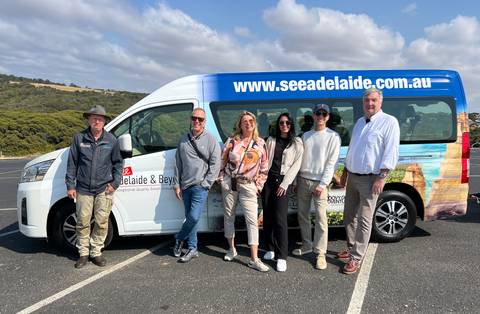       Tour group poses beside branded minibus on a coastal Australian road
  