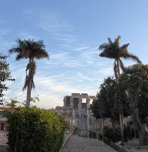       Distant Kom Ombo Temple framed by tall palm trees under streaky clouds.
  