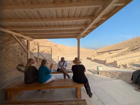       Small tour group sitting with guide under a wooden shelter in a desert valley setting.
  