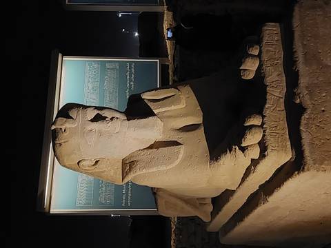       Close-up of a stone sphinx statue lit by spotlights in front of an information panel at night.
  