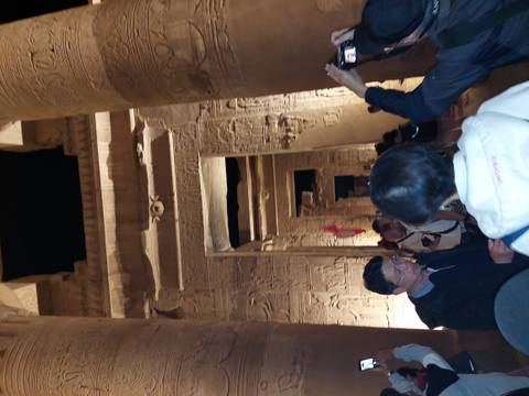       Night tour group walks through towering hieroglyph-covered columns inside a dimly lit temple.
  