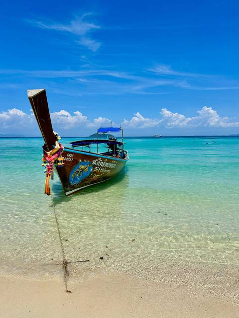       Traditional long-tail wooden boat decorated with flowers floating on clear turquoise water under a bright blue sky.
  