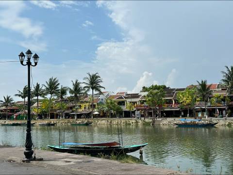       Charming riverside lined with palm trees and traditional shophouses reflected in calm water under a blue sky.
  