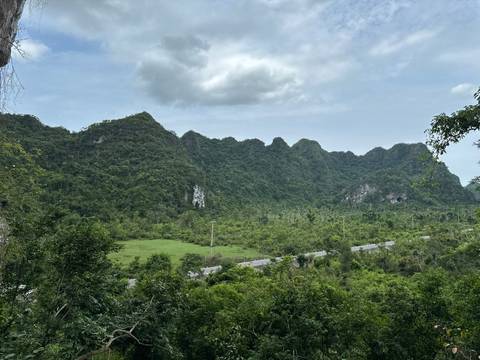       Dense green karst hills rising behind a valley road under a partly cloudy sky.
  