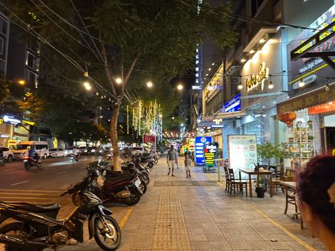       Vibrant, well-lit city street at night with motorbikes parked along the sidewalk and strings of fairy lights overhead.
  