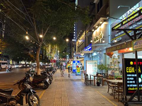       Nighttime city sidewalk scene with restaurants, neon signs, parked scooters and decorative string lights.
  