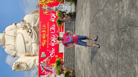       Traveller poses before a giant smiling Buddha statue and vibrant Tet decorations.
  
