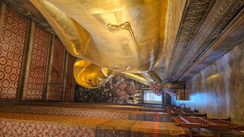       Golden reclining Buddha statue inside an ornately decorated temple hall with patterned ceiling and walls.
  