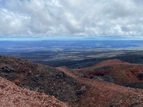       Expansive volcanic landscape with red and black lava fields stretching to distant horizon under cloud cover.
  