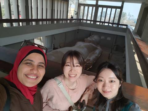       Smiling trio posing inside museum balcony overlooking colossal reclining statue of Ramses II.
  