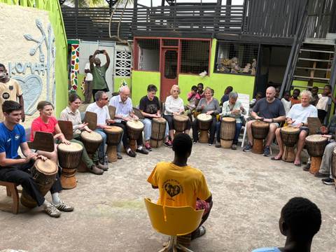       Large group seated in circle playing djembes during interactive drumming workshop.
  