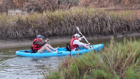       Two people paddle blue kayak through narrow marsh channel with tall grasses.
  