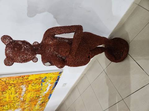       Indoor gallery shot of a woven wire sculpture of a pregnant female figure standing on a plinth against a white wall with modern art in the background.
  