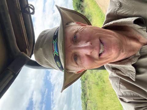       Selfie of a man wearing a safari hat in an open vehicle with savanna backdrop.
  