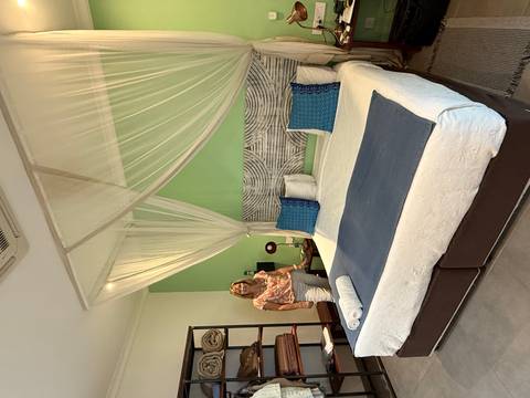       Neat hotel room with a king bed, mosquito net canopy and a smiling guest standing beside the bed.
  