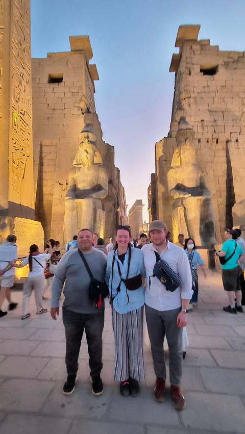       Travelers posing between colossal statues at the illuminated entrance of Luxor Temple at dusk.
  