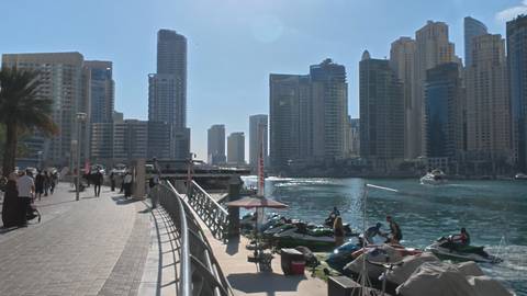       Pedestrians stroll the sunny promenade beside turquoise waters and high-rise towers of Dubai Marina.
  