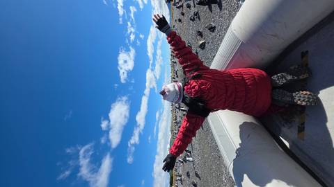       Kneeling traveler in a red jacket on an inflatable boat surrounded by hundreds of penguins under a bright blue sky.
  
