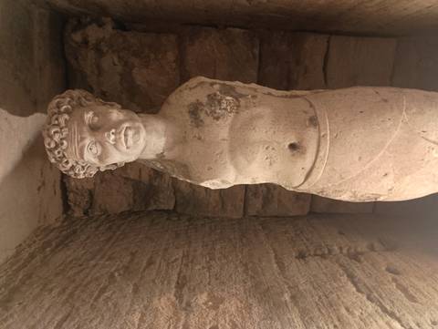       Illuminated marble torso statue displayed in a stone-walled underground chamber.
  