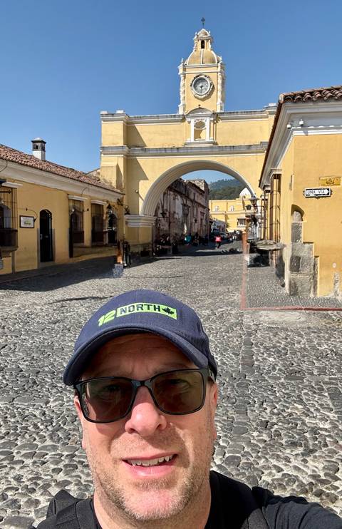       Partial selfie with the yellow Santa Catalina Arch spanning a cobbled street in Antigua
  