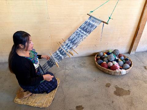       Indigenous woman weaving with a backstrap loom beside a basket of colourful yarn balls
  