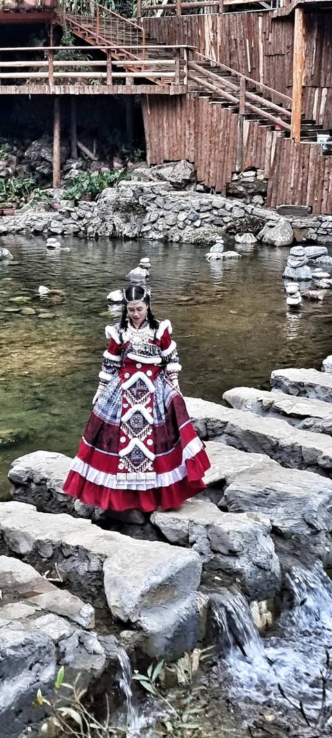       Young woman wearing an ornate traditional red and white costume standing on stone steps beside a calm stream.
  