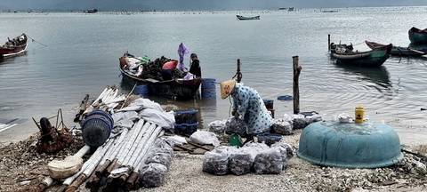       Local fishers sorting gear beside a tranquil lagoon with small wooden boats and bundled supplies on the shore.
  
