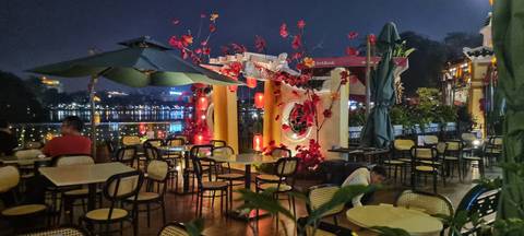       Riverside night cafe with red lanterns and empty tables overlooking city lights on the water.
  
