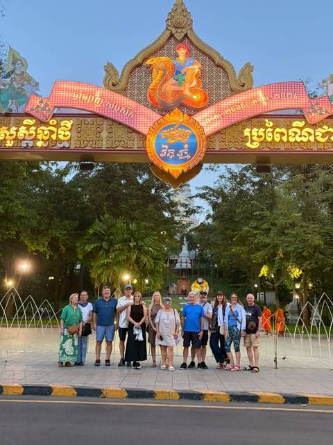       Tour group posing under an ornately carved entrance gate with trees and a stupa visible beyond
  