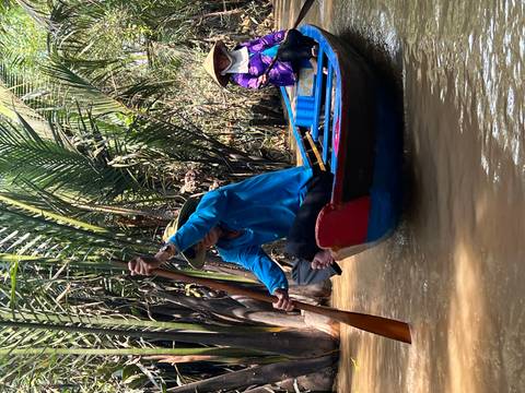       Local guide paddling a narrow wooden canoe through a palm-lined muddy canal with passenger aboard
  