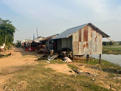       Rustic corrugated-iron stilt houses line a dusty rural track beside a small pond and open fields
  