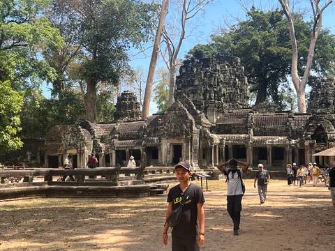       Visitors stroll past sprawling tree-root-covered ruins of Angkor complex dappled in midday sun
  